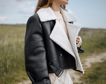 Könnte beinhalten: Schwarze Lederjacke mit weißem Shearling-Kragen und -Futter. Die Jacke hat ein kurzes Design mit Schnallenverschluss an Taille und Manschetten. Das Model trägt eine Sonnenbrille und einen cremefarbenen Pullover und ein Kleid.