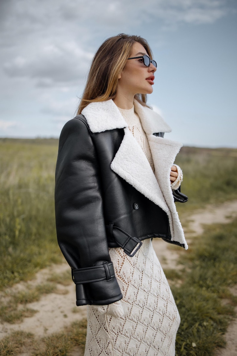 May include: A black leather jacket with a white shearling collar and lining. The jacket has a cropped design with a buckle closure at the waist and cuffs. The model is wearing sunglasses and a cream-colored sweater and dress.