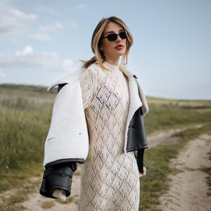 May include: A woman wearing a cream-colored, diamond-patterned knit dress and black sunglasses. A black leather jacket with a white shearling lining is draped over the shoulders. The background is a dirt path and grassy field.