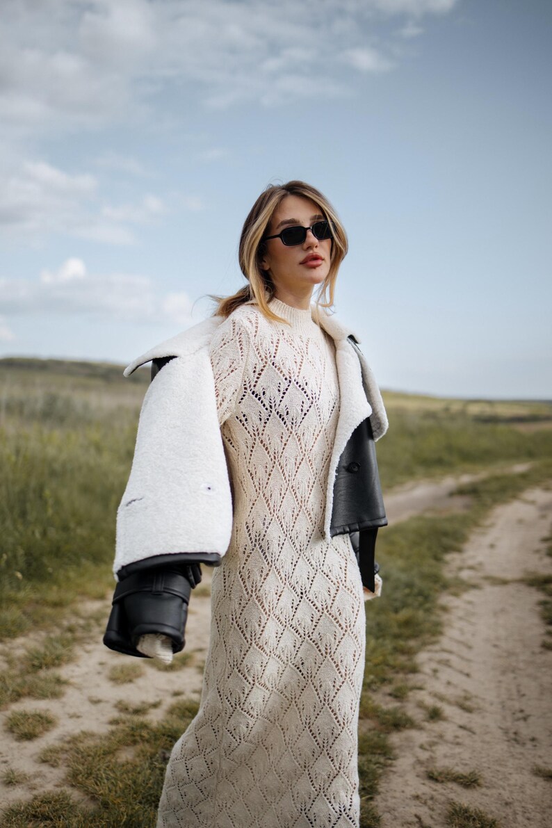 May include: A woman wearing a cream-colored, diamond-patterned knit dress and black sunglasses. A black leather jacket with a white shearling lining is draped over the shoulders. The background is a dirt path and grassy field.