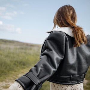 May include: A black leather jacket with a white shearling collar and cuffs. The jacket has a cropped design and is worn over a cream-colored, open-knit dress. The background features a field of green grass and a blue sky.
