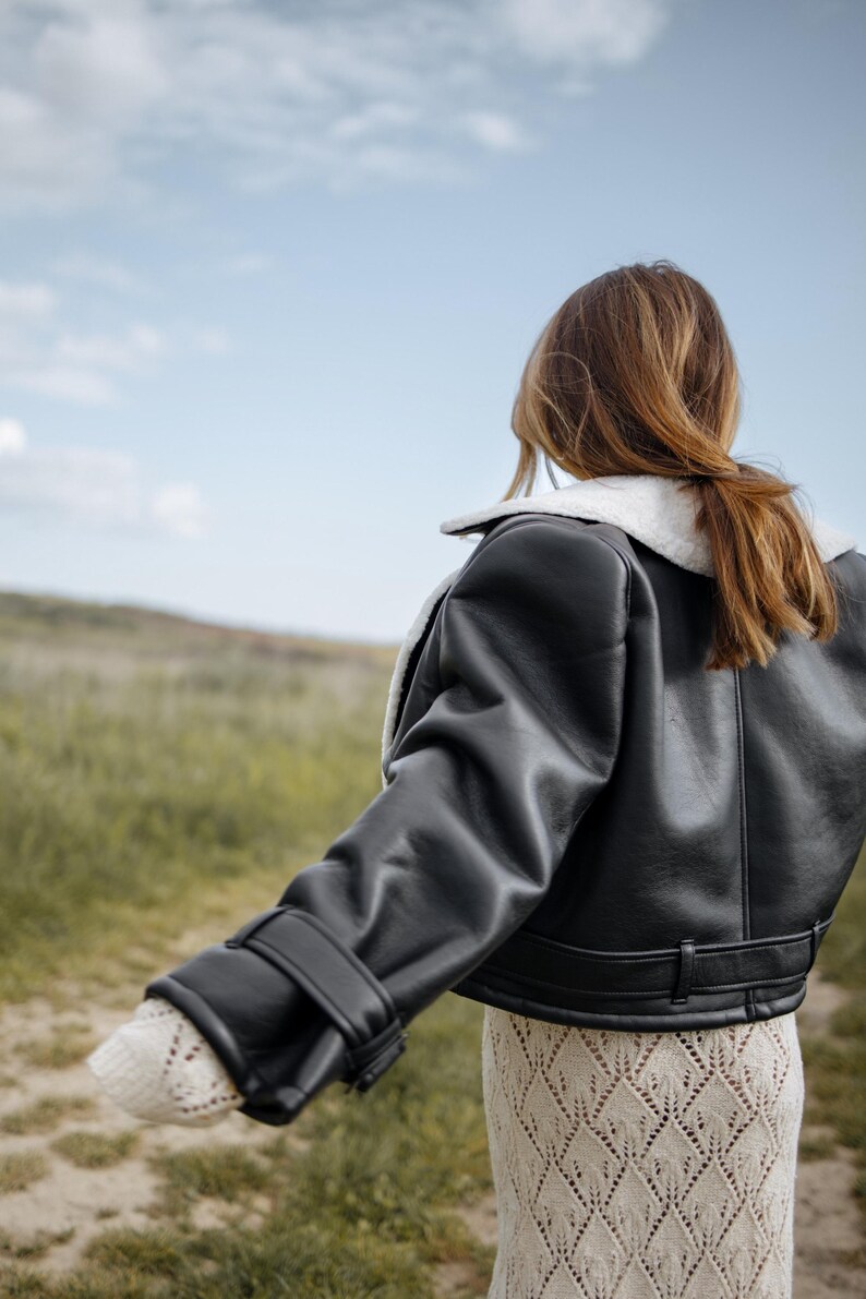 May include: A black leather jacket with a white shearling collar and cuffs. The jacket has a cropped design and is worn over a cream-colored, open-knit dress. The background features a field of green grass and a blue sky.