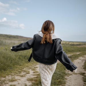 May include: A black leather jacket with a white shearling collar and cuffs. The jacket is worn over a cream-colored, open-knit dress. The person is walking on a dirt path through a field of green grass under a blue sky.