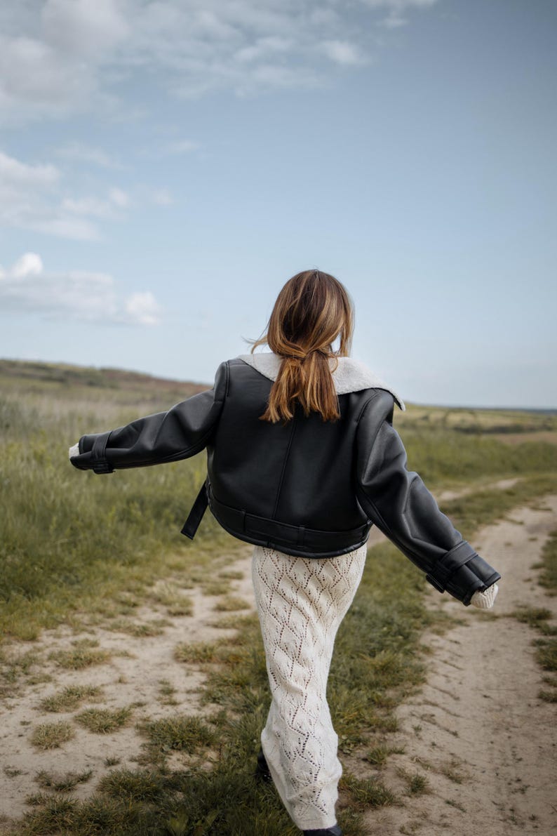 May include: A black leather jacket with a white shearling collar and cuffs. The jacket is worn over a cream-colored, open-knit dress. The person is walking on a dirt path through a field of green grass under a blue sky.
