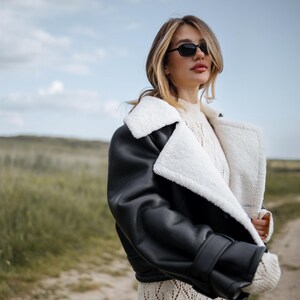 May include: A black leather jacket with a white shearling lining and collar. The jacket is worn over a cream-colored sweater and a patterned dress. The person is wearing black sunglasses and standing outdoors on a cloudy day.