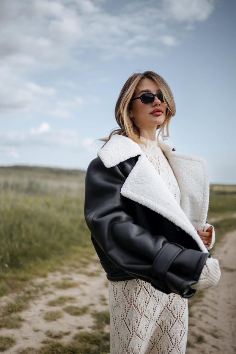 May include: A black leather jacket with a white shearling lining and collar. The jacket is worn over a cream-colored sweater and a patterned dress. The person is wearing black sunglasses and standing outdoors on a cloudy day.