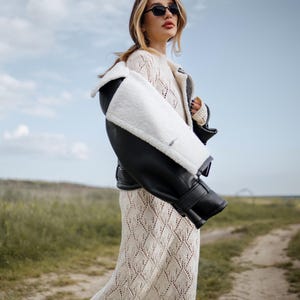 May include: A woman wearing a cream-colored, diamond-patterned dress and a black and white shearling jacket. She is also wearing black sunglasses. The background features a dirt path and a field under a cloudy sky.
