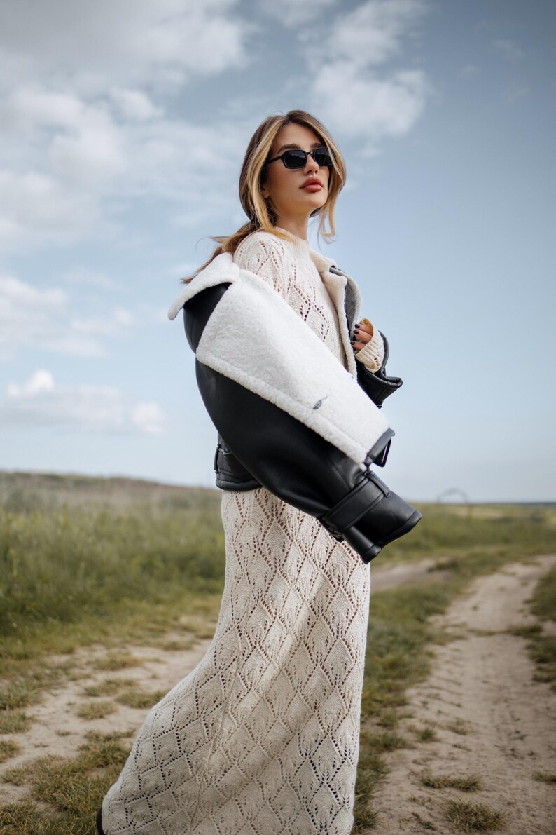 May include: A woman wearing a cream-colored, diamond-patterned dress and a black and white shearling jacket. She is also wearing black sunglasses. The background features a dirt path and a field under a cloudy sky.