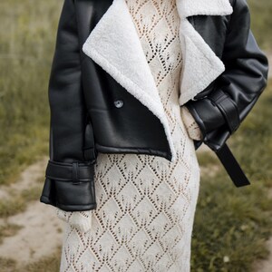 May include: A black faux leather jacket with a white shearling collar and cuffs. The jacket has a double-breasted closure and a belt detail. The person is wearing a cream-colored, open-knit dress.