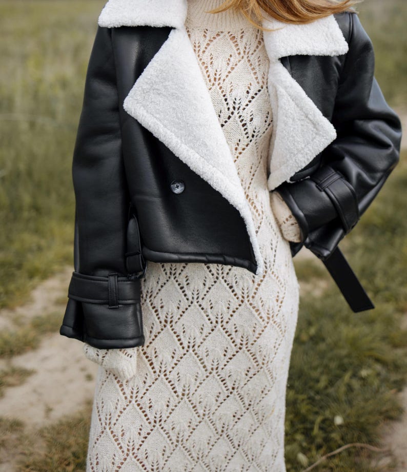 May include: A black faux leather jacket with a white shearling collar and cuffs. The jacket has a double-breasted closure and a belt detail. The person is wearing a cream-colored, open-knit dress.