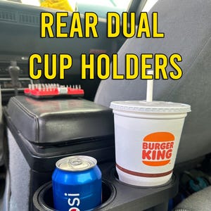 May include: A black plastic dual cup holder for a car. It has two cup holders, one holding a can of soda and the other holding a Burger King cup. The cup holder is mounted on the back of the centre console.