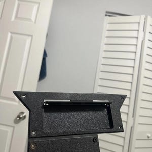 Peut inclure: Un composant en plastique noir avec une surface texturée. Le composant comporte une ouverture rectangulaire et plusieurs trous de montage. Une barre métallique est visible à l'intérieur de l'ouverture. Le composant semble faire partie d'un assemblage plus grand.