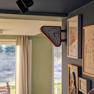 May include: A unique, triangular wall clock with a light wood frame and a white clock face. The clock is mounted on a corner wall, with a view of a window and other framed artwork. The clock's hands indicate the time.