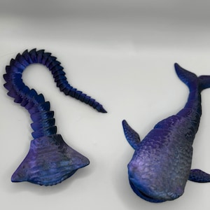 May include: Two whimsical, purple and blue sculptures. One resembles a whale with fins and a tail. The other is a curved, segmented form with a flared base. Both have a textured surface and are set against a white background.