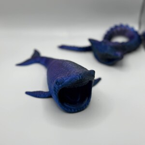 May include: Two blue whale figurines with open mouths and detailed textures. The whales are painted in shades of blue and purple, with visible teeth. One whale is in focus, while the other is slightly blurred in the background.
