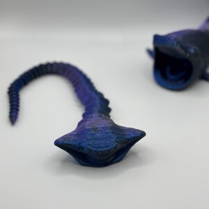 May include: Two 3D-printed, stylized whale shark models in shades of blue and purple. One model is complete with an open mouth, while the other is a detached tail section. The models have textured surfaces and are set against a white background.