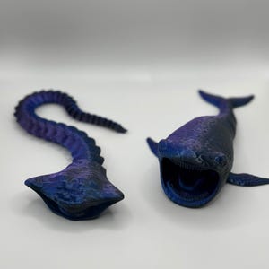 May include: Two blue and purple 3D-printed sculptures. One is a whale with an open mouth, the other is a curved, segmented form. The sculptures have a textured surface and are set against a white background.