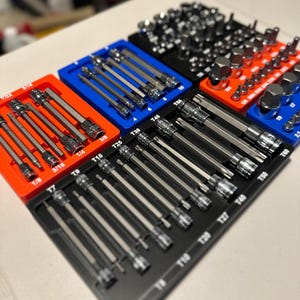 May include: A set of various metal bits and sockets organized in colorful trays. The trays are orange, blue, and black, with sizes labeled in inches. The tools are arranged neatly, showcasing a variety of sizes and types for mechanical work.