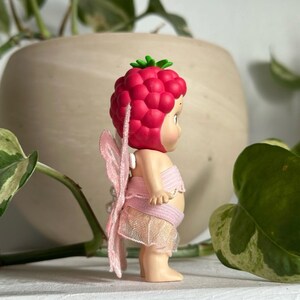 K&ouml;nnte beinhalten: Kleine Fee-Figur mit himbeerf&ouml;rmiger Kopfbedeckung und rosa Fl&uuml;geln. Die Fee tr&auml;gt ein rosa Oberteil und einen Rock. Die Figur steht auf einer wei&szlig;en Oberfl&auml;che, mit einem verschwommenen Hintergrund aus Gr&uuml;n und einem beigen Topf.