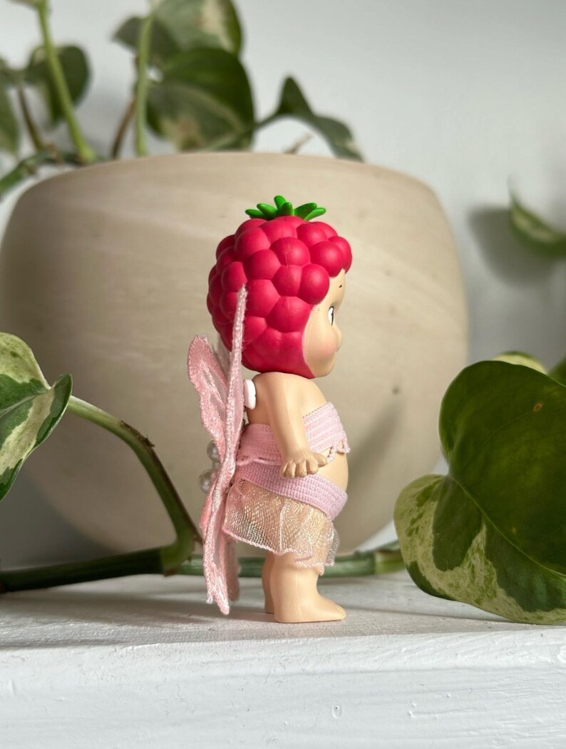 K&ouml;nnte beinhalten: Kleine Fee-Figur mit himbeerf&ouml;rmiger Kopfbedeckung und rosa Fl&uuml;geln. Die Fee tr&auml;gt ein rosa Oberteil und einen Rock. Die Figur steht auf einer wei&szlig;en Oberfl&auml;che, mit einem verschwommenen Hintergrund aus Gr&uuml;n und einem beigen Topf.