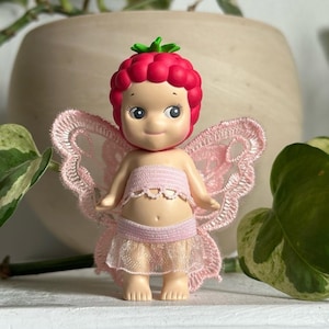 K&ouml;nnte beinhalten: Eine skurrile Fee-Figur mit einem himbeerf&ouml;rmigen Kopf und einem l&auml;chelnden Gesicht. Die Fee tr&auml;gt ein rosa Oberteil, einen geschichteten Rock und zarte Spitzenfl&uuml;gel. Ein Sammlerst&uuml;ck.