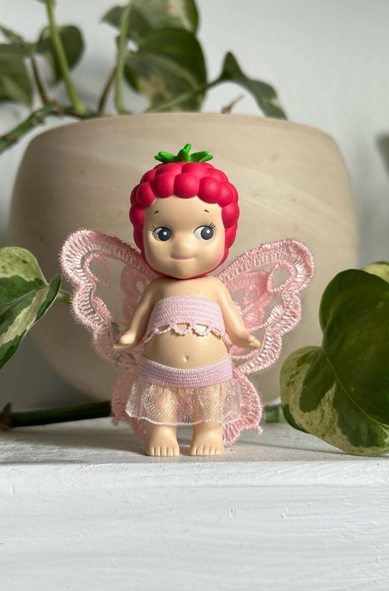 K&ouml;nnte beinhalten: Eine skurrile Fee-Figur mit einem himbeerf&ouml;rmigen Kopf und einem l&auml;chelnden Gesicht. Die Fee tr&auml;gt ein rosa Oberteil, einen geschichteten Rock und zarte Spitzenfl&uuml;gel. Ein Sammlerst&uuml;ck.