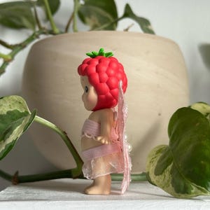 K&ouml;nnte beinhalten: Kleine Figur einer Babyfee mit rotem Himbeerkopfschmuck und rosa Fl&uuml;geln. Die Fee tr&auml;gt ein rosa Oberteil und einen Rock. Die Figur steht auf einer wei&szlig;en Oberfl&auml;che mit gr&uuml;nen Pflanzen im Hintergrund.