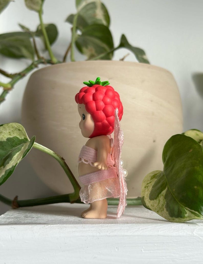 K&ouml;nnte beinhalten: Kleine Figur einer Babyfee mit rotem Himbeerkopfschmuck und rosa Fl&uuml;geln. Die Fee tr&auml;gt ein rosa Oberteil und einen Rock. Die Figur steht auf einer wei&szlig;en Oberfl&auml;che mit gr&uuml;nen Pflanzen im Hintergrund.