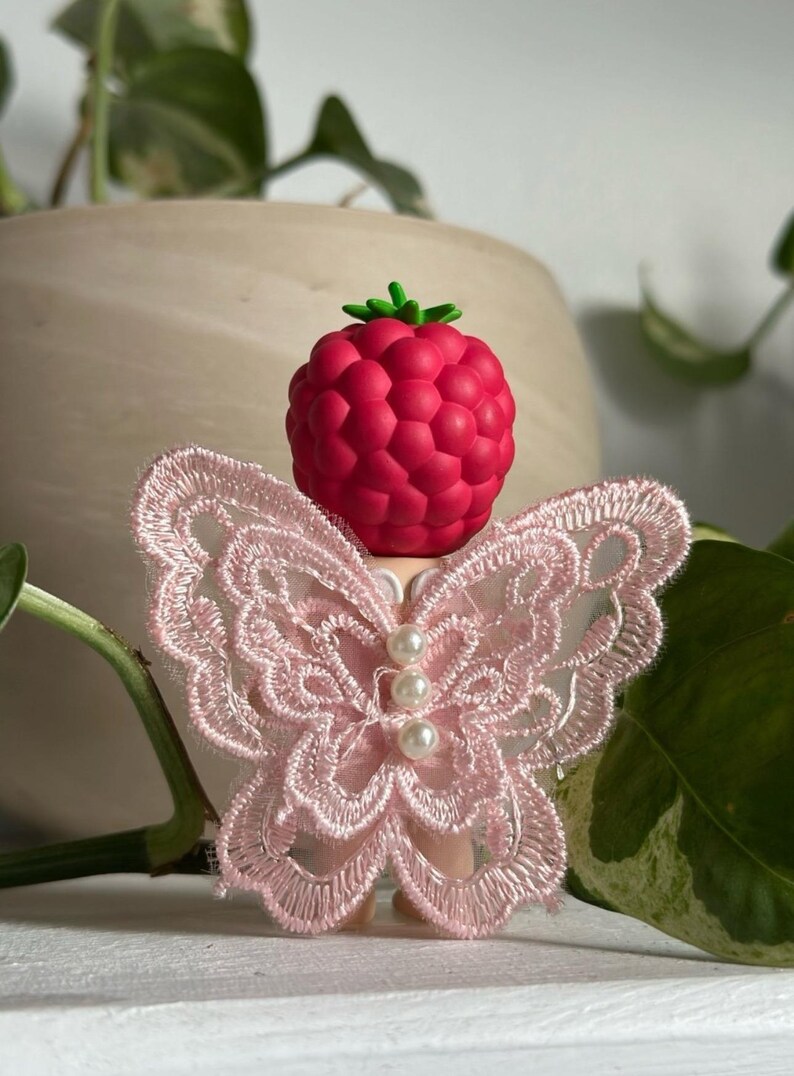 K&ouml;nnte beinhalten: Ein dekorativer Flaschenverschluss mit einem Himbeerdesign und einem gr&uuml;nen Stiel. Er verf&uuml;gt &uuml;ber rosa Spitzen-Schmetterlingsfl&uuml;gel und drei Perlenakzente. Dieser Verschluss soll jeder Flasche einen Hauch von Eleganz verleihen.