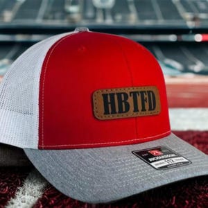 May include: Red and white trucker hat with a grey brim. The hat features a brown leather patch with the letters "HBTFD" and a Richardson 112 tag.