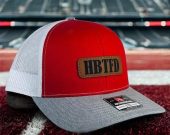 May include: Red and white trucker hat with a gray brim. The hat features a brown leather patch with the letters "HBTFD" and a Richardson 112 tag.