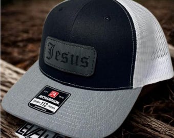 May include: A black, gray, and white trucker hat with the word "Jesus" on a leather patch. The hat has a gray brim and a white mesh back. A "Richardson" tag is visible.