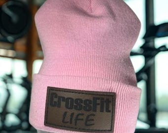 May include: A light pink beanie with a brown rectangular patch. The patch features the words "CrossFit LIFE" in a bold, dark font. The beanie is made of a knitted material.