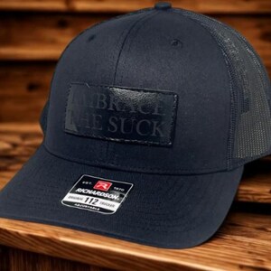 May include: Navy blue trucker hat with a mesh back and a curved brim. The hat features a rectangular patch with the words "EMBRACE THE SUCK" embossed on it. The hat is adjustable and has a Richardson tag.