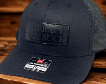 May include: Navy blue trucker hat with a mesh back and a curved brim. The hat features a rectangular patch with the words "EMBRACE THE SUCK" embossed on it. The hat is adjustable and has a Richardson tag.