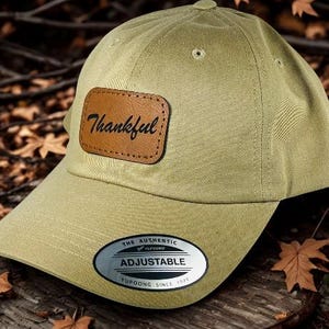 May include: A khaki-colored baseball cap with a curved brim. A brown leather patch on the front displays the word "Thankful" in cursive. The cap has an adjustable strap and a Yupoong tag.