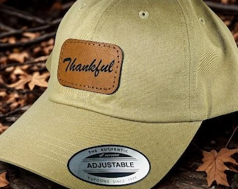 May include: A khaki-colored baseball cap with a curved brim. A brown leather patch on the front displays the word "Thankful" in cursive. The cap has an adjustable strap and a Yupoong tag.