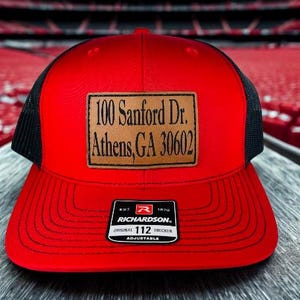 May include: Red trucker hat with a black mesh back and a brown leather patch. The patch displays the address "100 Sanford Dr. Athens, GA 30602". The hat has a Richardson 112 Trucker adjustable tag.