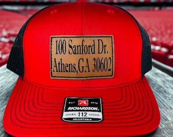 May include: Red trucker hat with a black mesh back and a brown leather patch. The patch displays the address "100 Sanford Dr. Athens, GA 30602". The hat has a Richardson 112 Trucker adjustable tag.