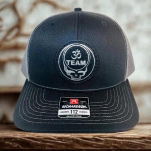 May include: A navy blue and grey trucker cap with a white embroidered patch featuring the Om symbol, the word "TEAM", and a skull design. The cap has a curved peak and an adjustable strap. The Richardson tag is visible.