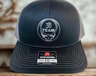 May include: A navy blue and gray trucker hat with a white embroidered patch featuring the Om symbol, the word "TEAM", and a skull design. The hat has a curved brim and an adjustable strap. The Richardson tag is visible.