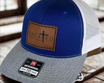 May include: A blue and gray trucker hat with a white mesh back. The hat features a brown leather patch with the word "CREATED" and a cross design. The hat is adjustable and has a Richardson 112 Trucker tag.