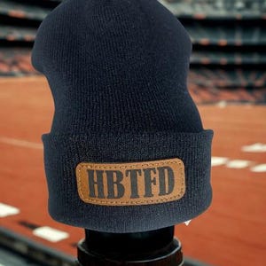 May include: A navy blue knit beanie with a brown leather patch. The patch features the letters "HBTFD" in a bold, sans-serif font. The beanie is displayed against a blurred background of a stadium.