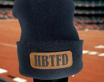 May include: A navy blue knit beanie with a brown leather patch. The patch features the letters "HBTFD" in a bold, sans-serif font. The beanie is displayed against a blurred background of a stadium.