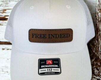 May include: White trucker hat with a mesh back and a curved brim. The hat features a brown leather patch with the text "FREE INDEED". A black and white Richardson 112 Trucker label is on the front.