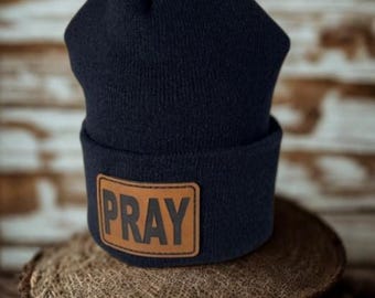 May include: A navy blue knit beanie with a brown leather patch that reads "PRAY" in black letters. The beanie is sitting on a round, woven surface.