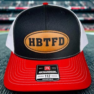 May include: A black, white, and red Richardson 112 trucker hat with the text "HBTFD" on a brown leather patch. The hat has a red brim and a white mesh back. The hat is adjustable.