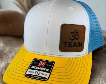 May include: A white, blue, and yellow trucker hat with a brown leather patch featuring the Om symbol and the word "TEAM." The hat has a curved brim and a mesh back. The label reads "RICHARDSON 112 TRUCKER ADJUSTABLE."