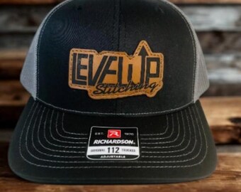 May include: Black and gray trucker hat with a curved brim and mesh back. The hat features a brown leather patch with the words "LEVEL UP Stitching" in a stylized font. The Richardson 112 hat is adjustable.