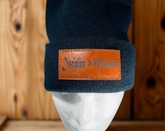 May include: A navy blue beanie with a brown leather patch. The patch features the text "Discipline > Motivation" in a stylized font. The beanie is displayed on a white mannequin head against a wooden background.